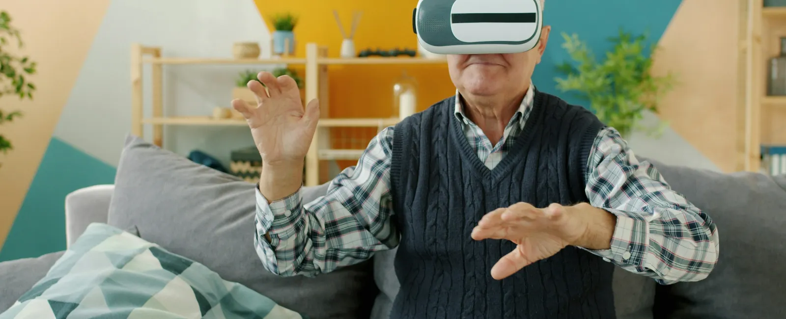 Elderly man wearing virtual reality headset interacting while sitting on a couch indoors with colorful background.