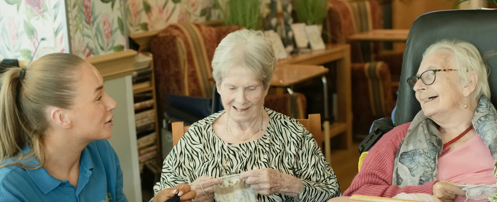 assisted living in atlanta: Young caregiver and two elderly women knitting together and smiling in a cozy room with art supplies on the table.