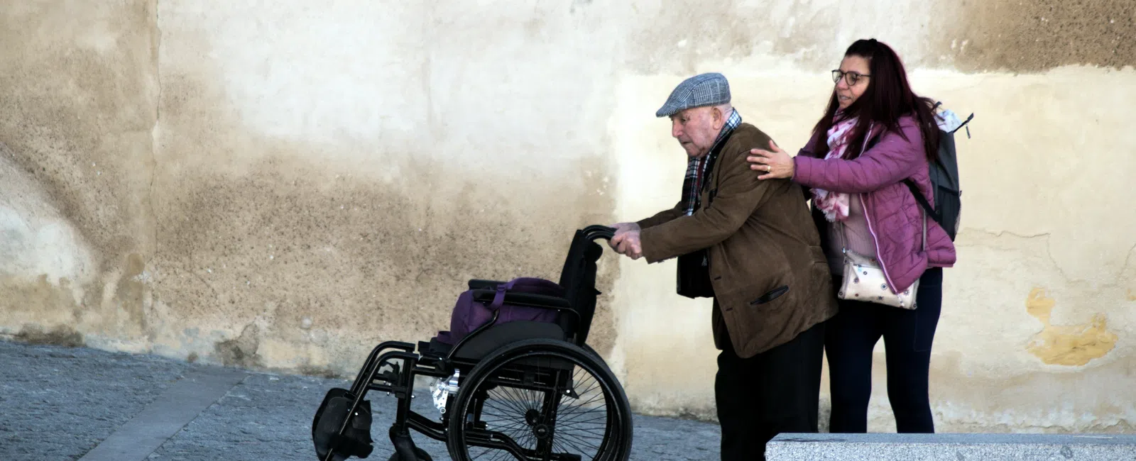 Young woman helping elderly man walk with wheelchair outside against textured wall