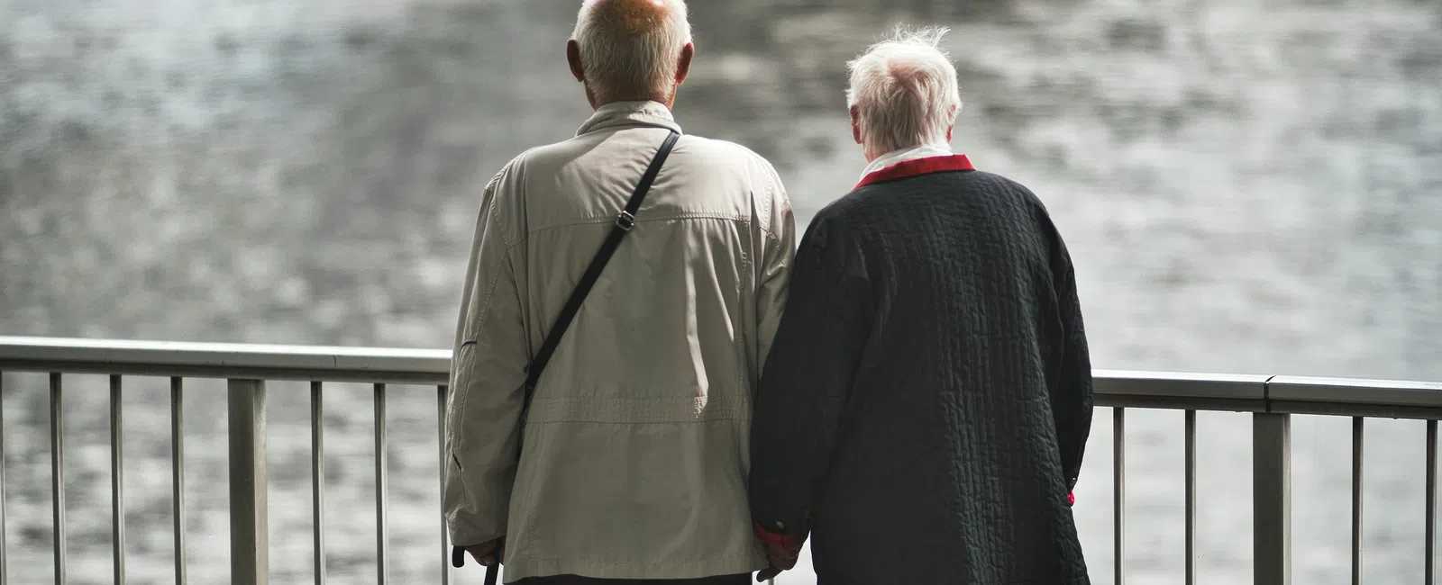 Elderly couple holding hands by a riverside railing under a large bridge on a cloudy day.