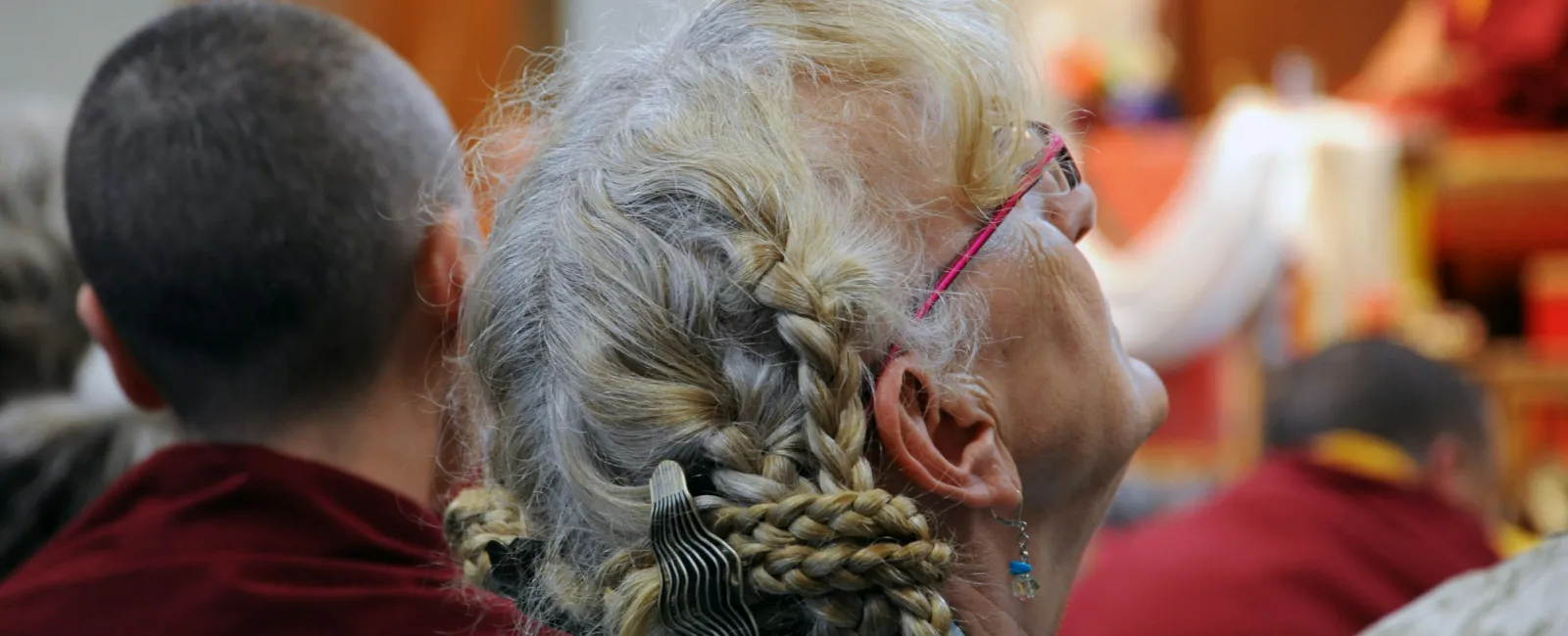 Elderly woman with braided gray hair and pink glasses listening intently during a gathering with others.