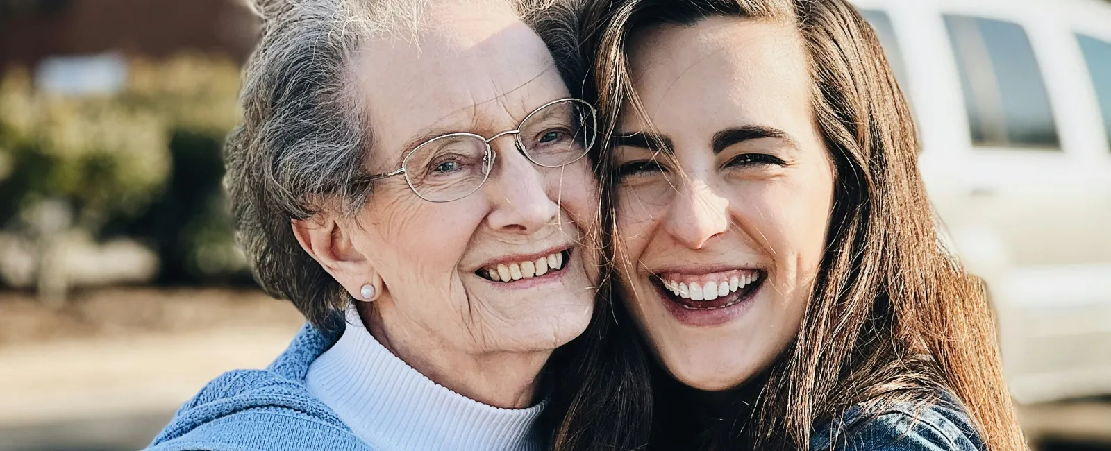 Smiling elderly woman and young woman hugging outdoors, both wearing sweaters and jackets in daylight.