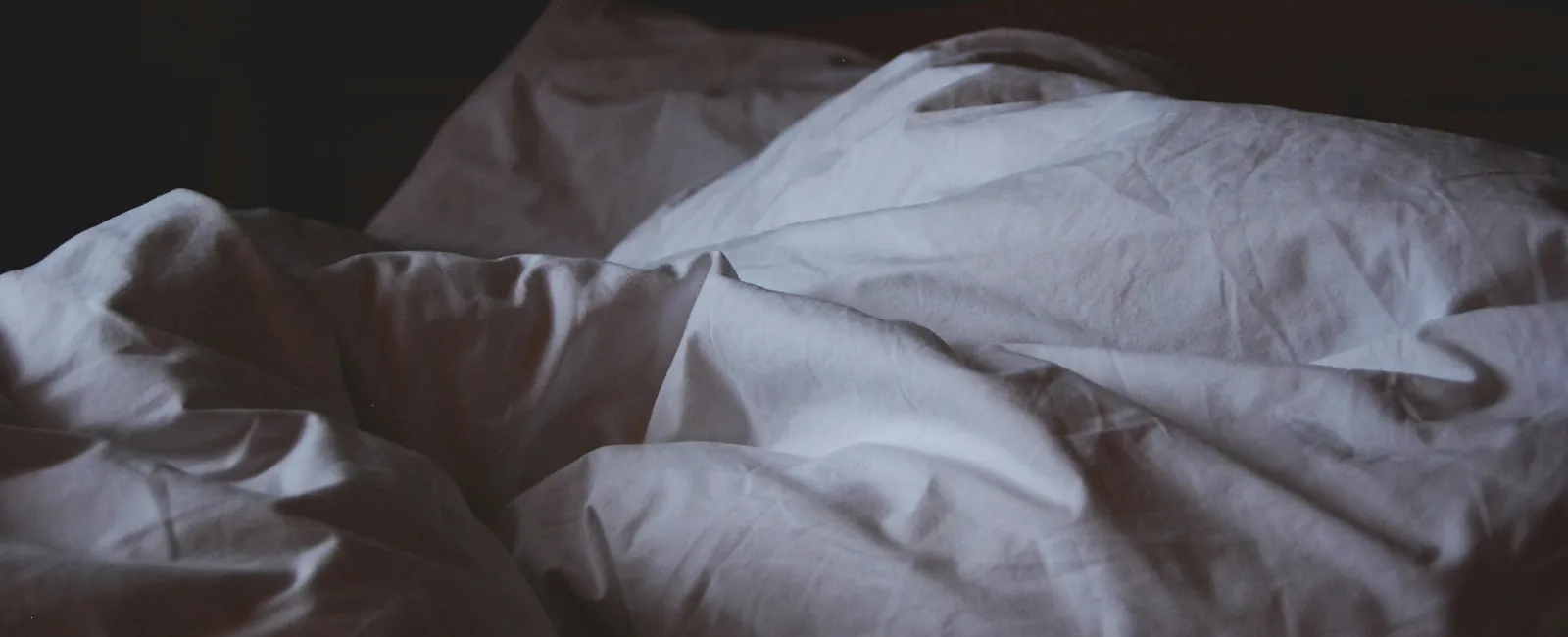 Crumpled white bed sheets and pillows in dim lighting on an unmade bed.