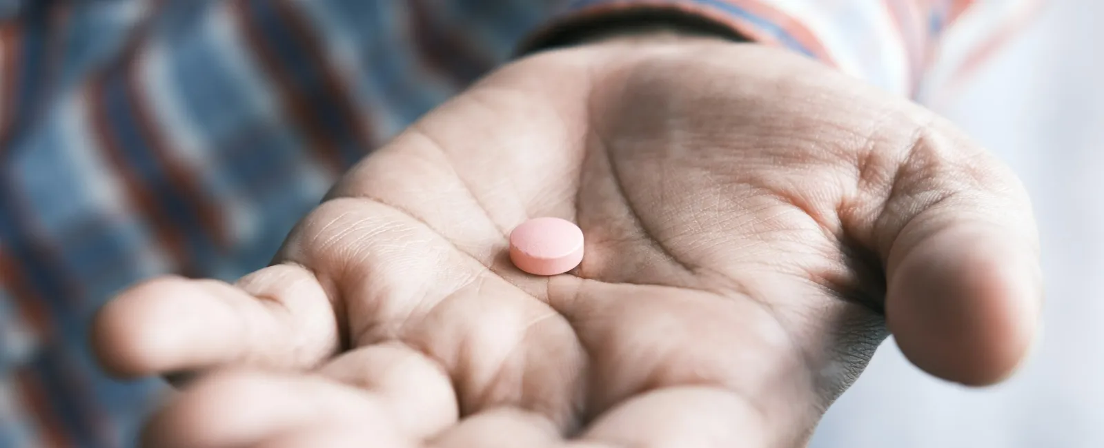 Close-up of an open hand holding a single pink pill with a blurred background of a striped shirt.