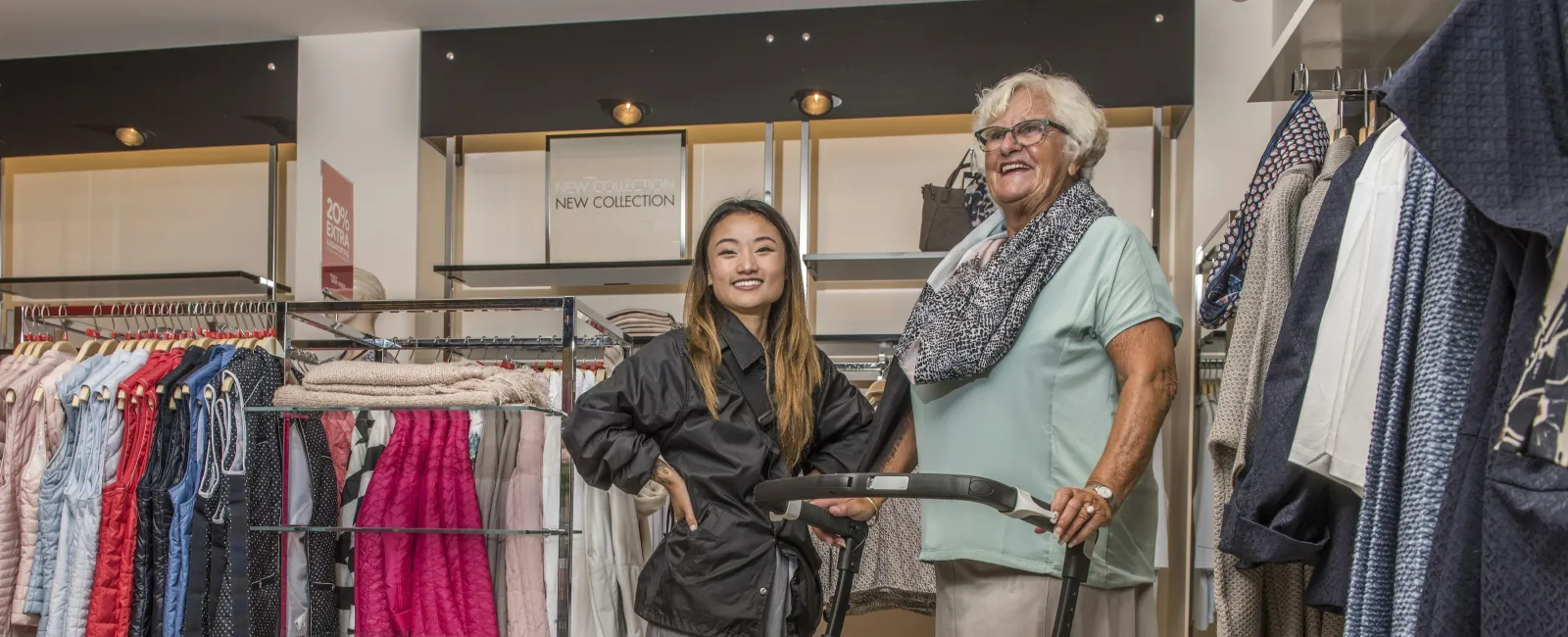 Young and elderly women smiling together in a clothing store among racks of colorful clothes.
