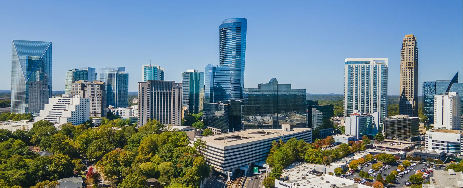 Aerial view of a modern city skyline with skyscrapers, busy highway, and lush green trees under clear blue sky.