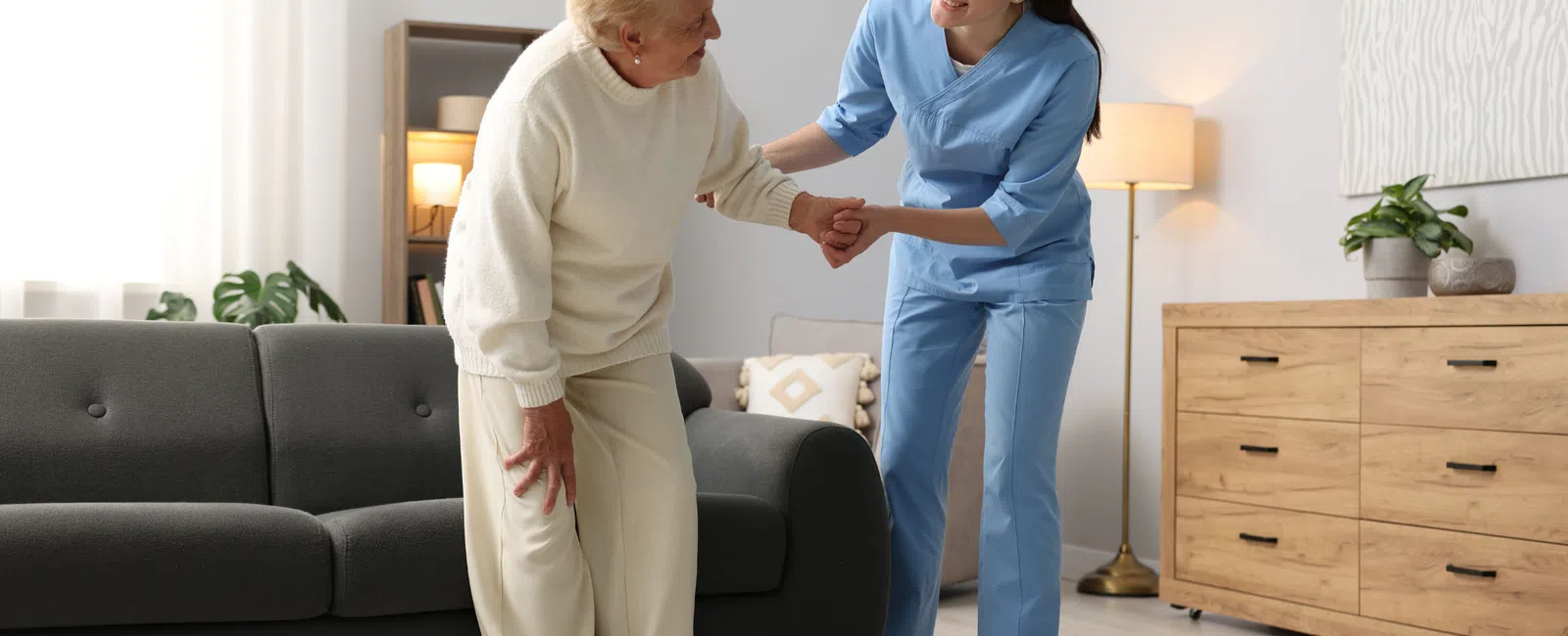 Nurse assisting elderly woman to stand from sofa in a bright, cozy living room with wooden furniture