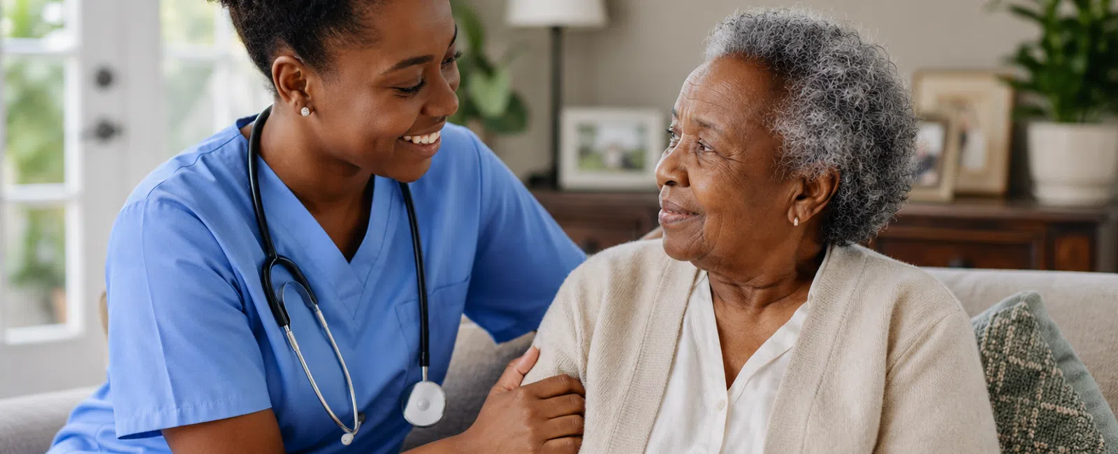 Smiling female nurse in blue scrubs comforting elderly woman sitting on sofa in cozy living room.