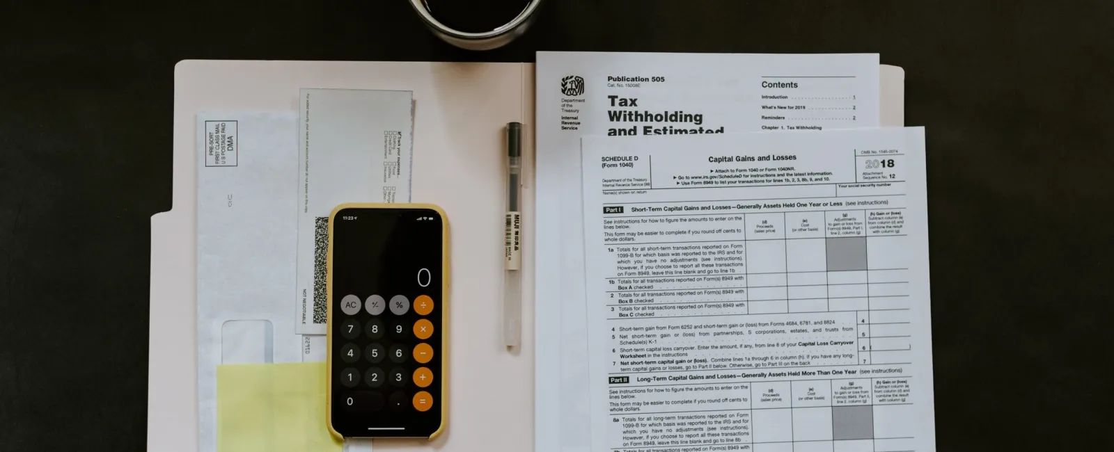 Smartphone calculator, tax forms, envelopes, pen, and coffee cup on dark desk for tax preparation.