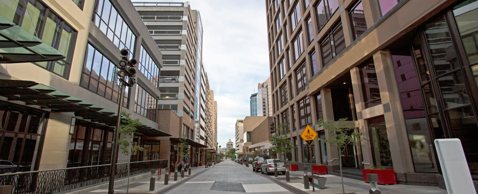 Empty urban street lined with modern office buildings and parked cars under a partly cloudy sky.