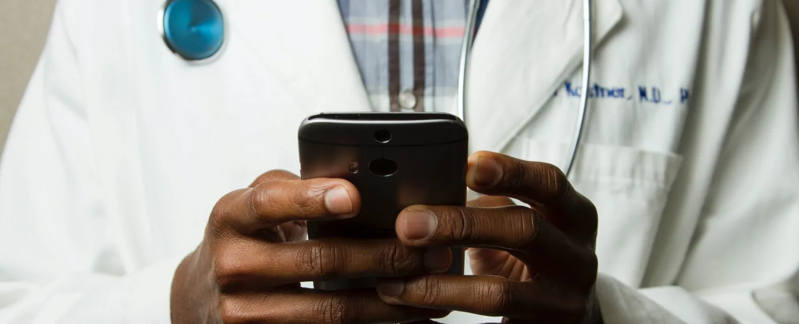 Doctor in white coat using smartphone with blue stethoscope around neck in clinical setting