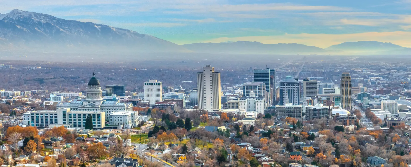 Panoramic view of a city skyline with autumn trees and mountains under a partly cloudy sky.