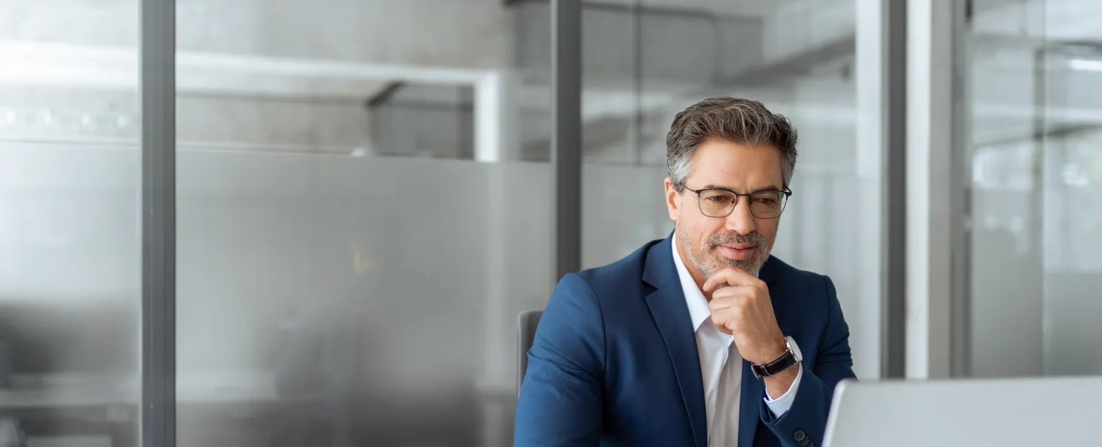 Professional man in a blue suit working thoughtfully on a laptop in a modern office setting.