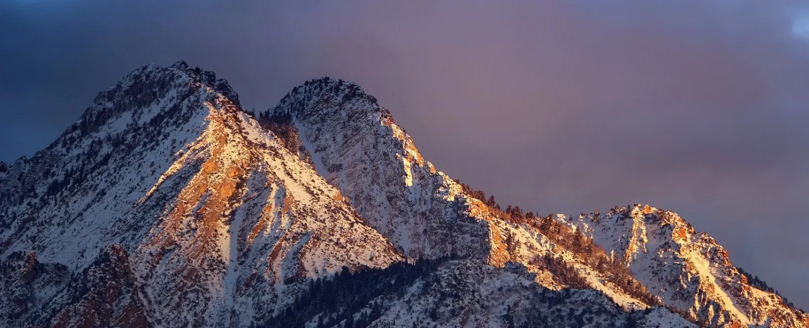 Snow-covered mountain peaks illuminated by golden sunlight under a dramatic cloudy sky at dusk.