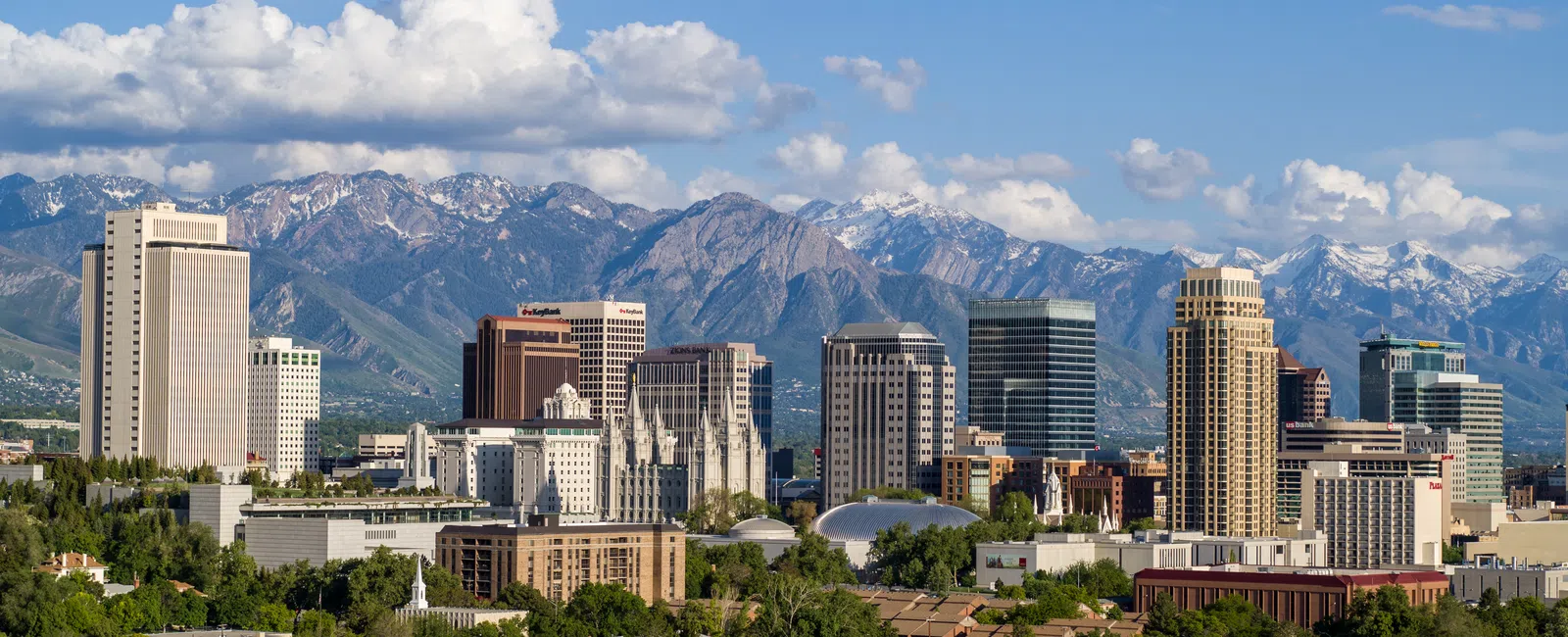 Salt Lake City skyline with modern buildings and snow-capped mountains under a partly cloudy blue sky