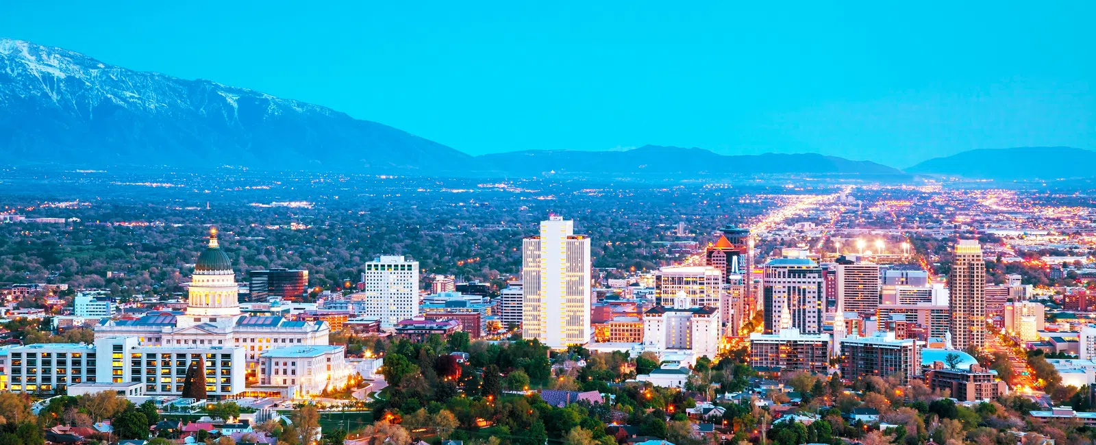 Evening skyline of Salt Lake City with lit buildings, green trees, suburbs, and snow-capped mountains in the background.