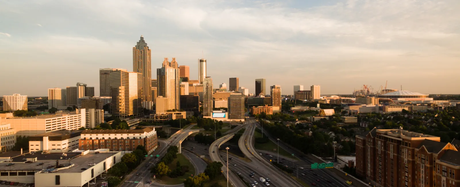 Aerial view of Atlanta skyline with highways, tall buildings, and a partly cloudy sky at sunset.