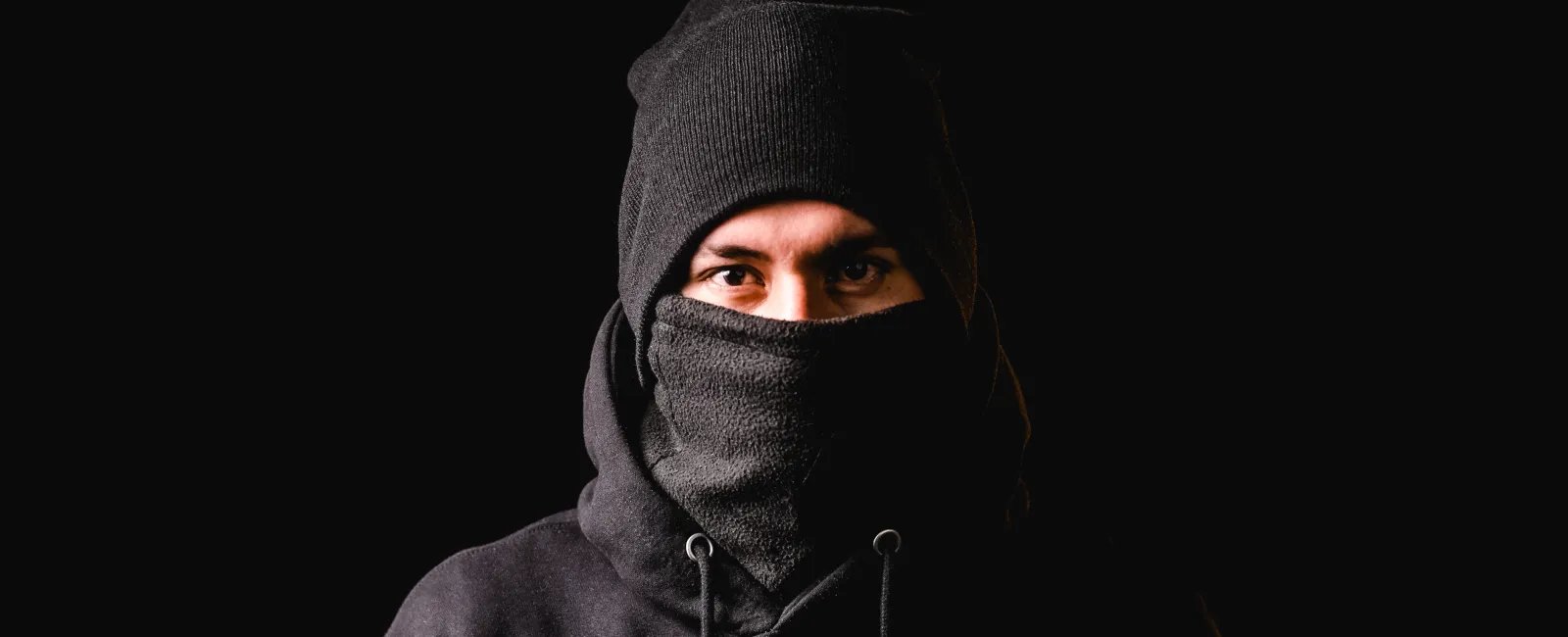 Man wearing black beanie and scarf covering his face against a dark background, showing intense eyes.