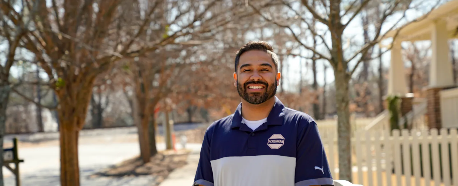 Smiling bearded man in blue-gray polo standing outdoors by a white picket fence on a sunny day.