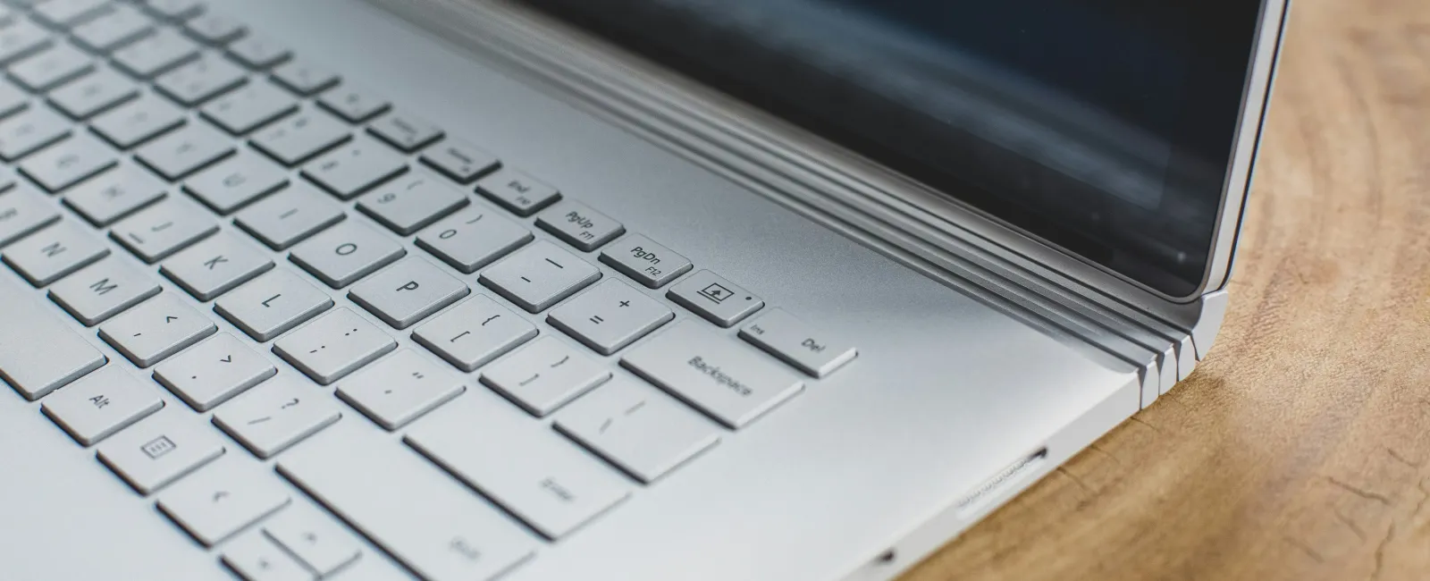 Close-up of a silver laptop keyboard and hinge on a wooden surface with a reflective screen.