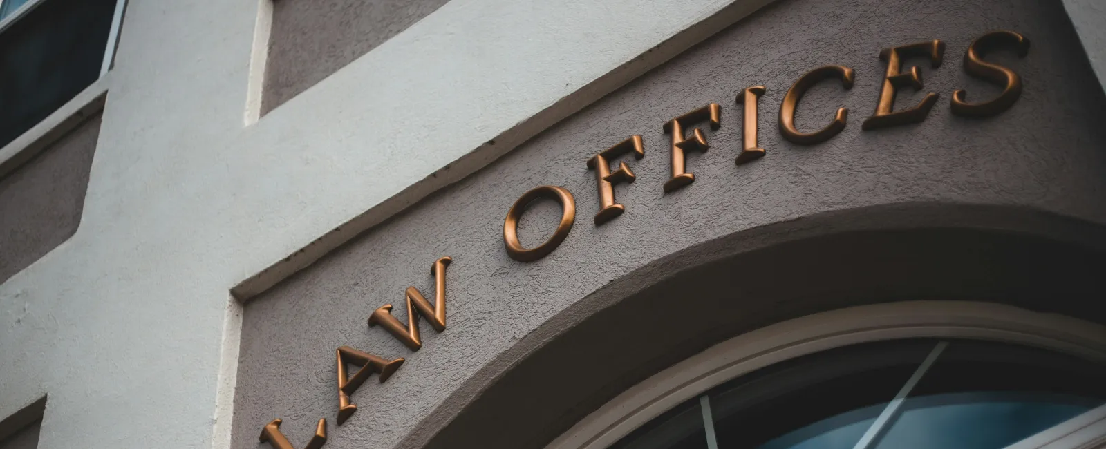 Brown lettering spelling LAW OFFICES on gray building facade above arched window with blue sky reflection.