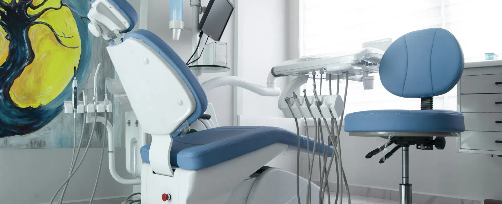 Modern dental clinic with blue dental chair, dentist stool, equipment, and colorful abstract wall art.