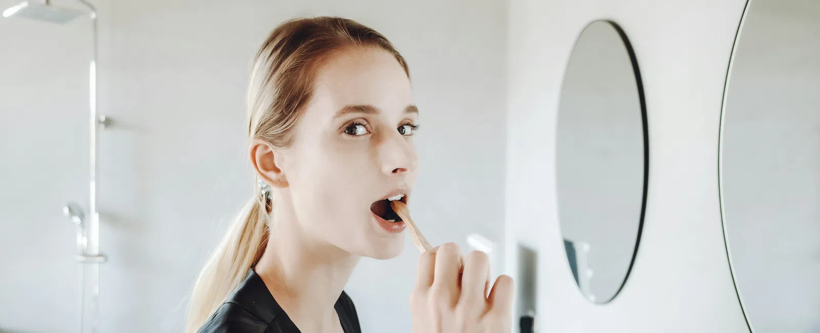 Young woman brushing teeth in modern bathroom with round mirror and shower in the background