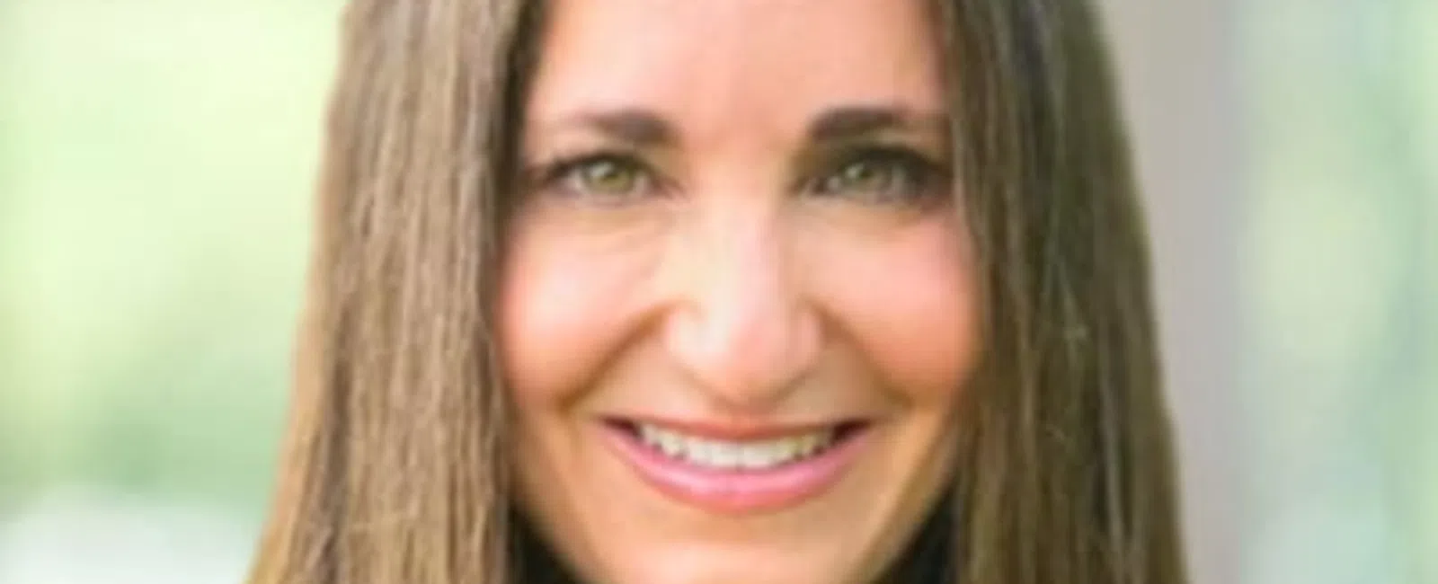 Smiling woman with long brown hair wearing a black top and white shirt, blurred outdoor background.