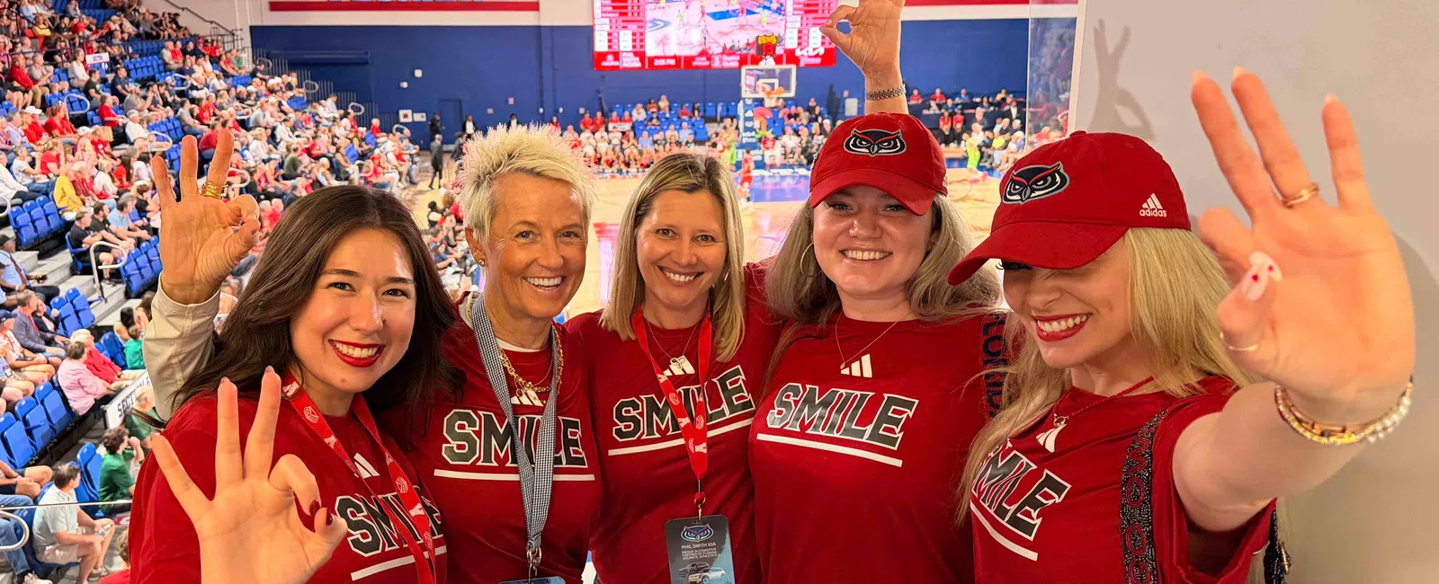 Five women in red SMILE shirts and hats smiling and making hand signs at a Florida Atlantic basketball game.