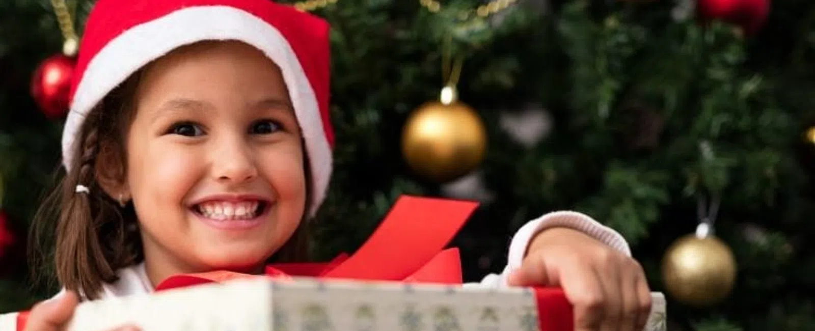 Smiling girl in Santa hat holding a wrapped Christmas present in front of a decorated tree with gold ornaments.
