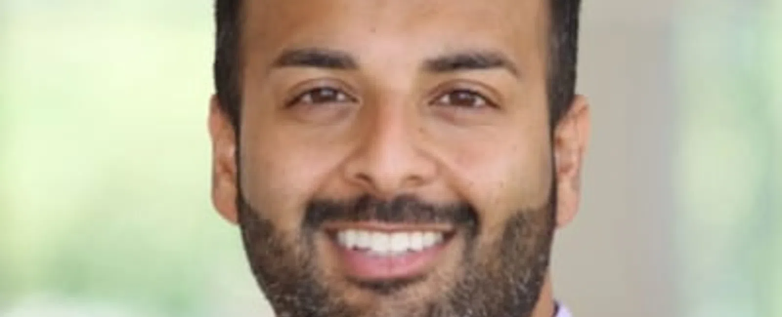 Smiling man with beard wearing a black suit, lavender shirt, and striped tie against a blurred background