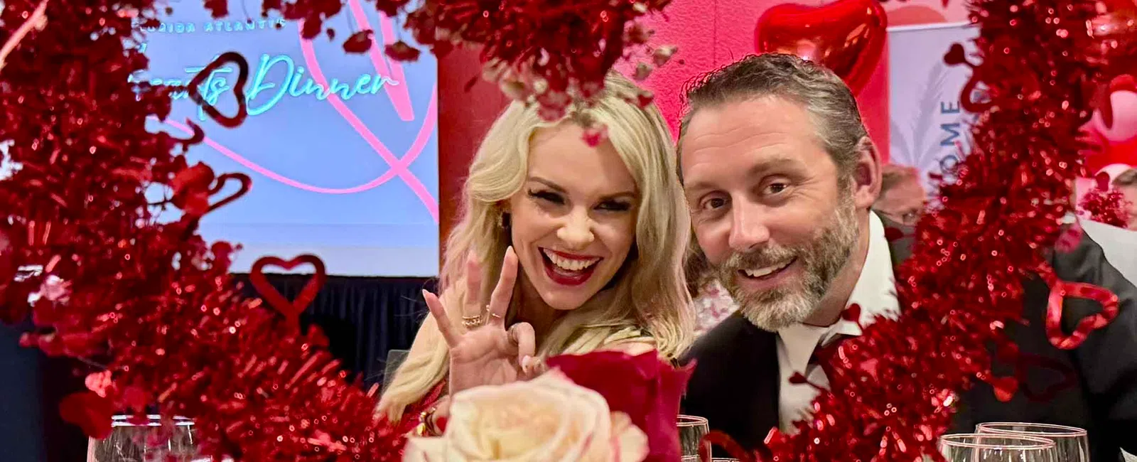 Smiling couple posing behind red heart-shaped decoration and roses at a romantic dinner event.