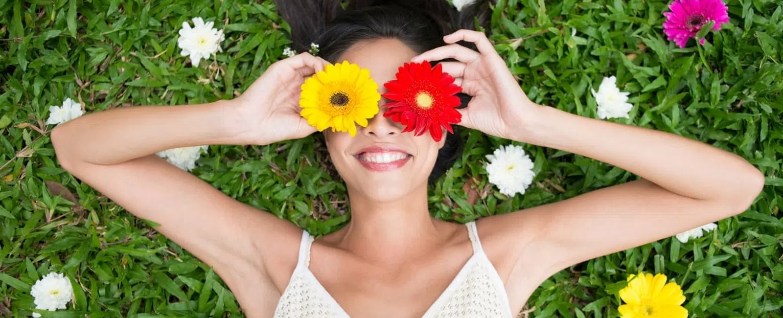 Smiling woman lying on grass holding yellow and red flowers over her eyes surrounded by white flowers.