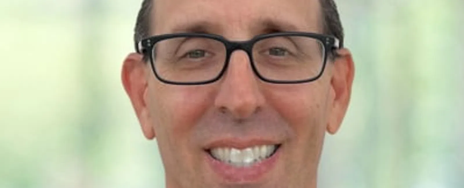 Smiling middle-aged man with glasses wearing black medical scrubs against a blurred background.