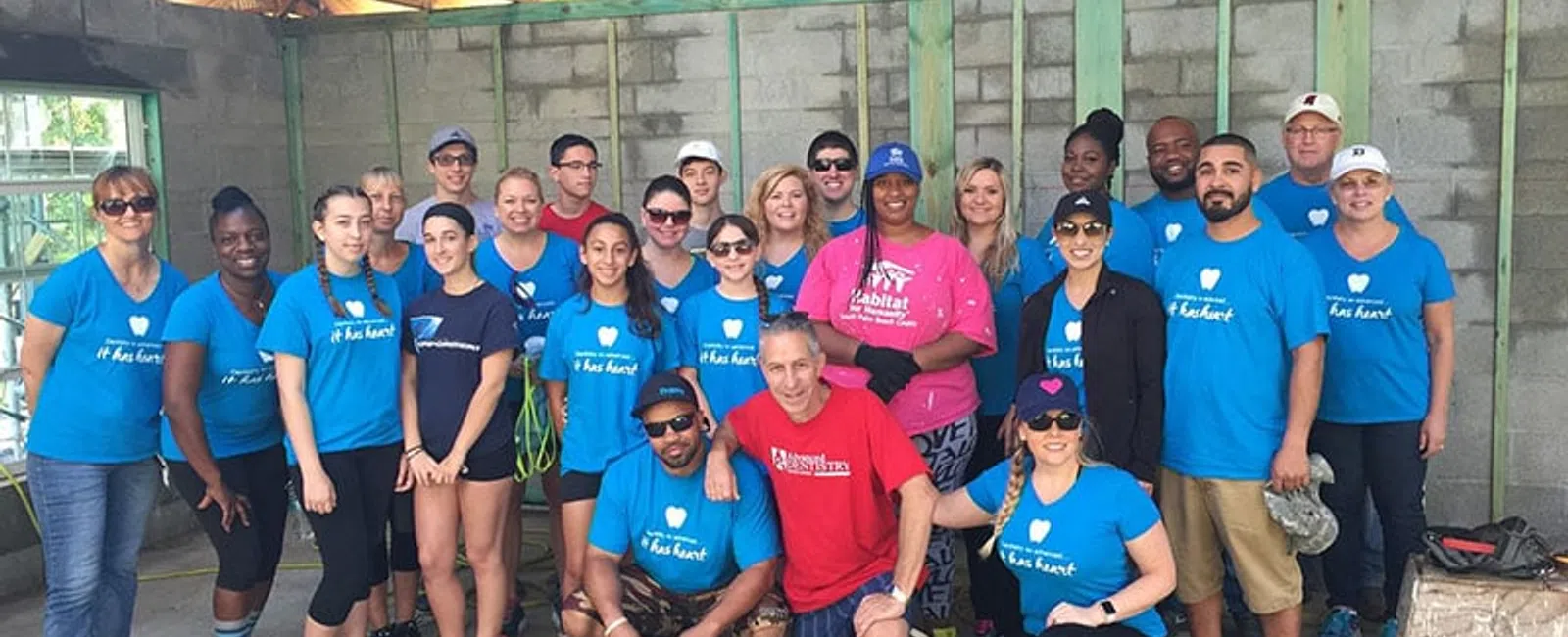 Group of diverse volunteers smiling inside a partially built house with wooden beams and concrete walls.