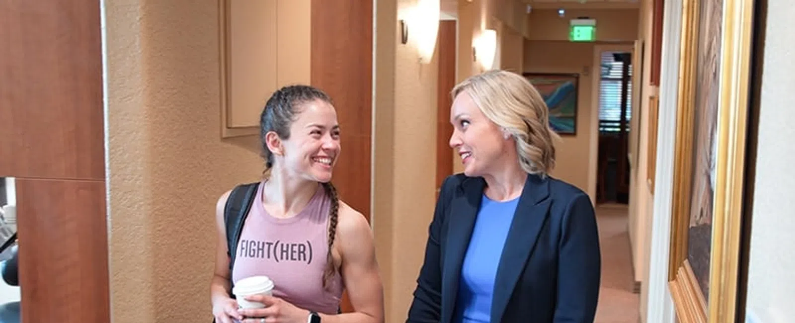 Two women smiling and talking while walking down a hallway in a professional and casual setting.