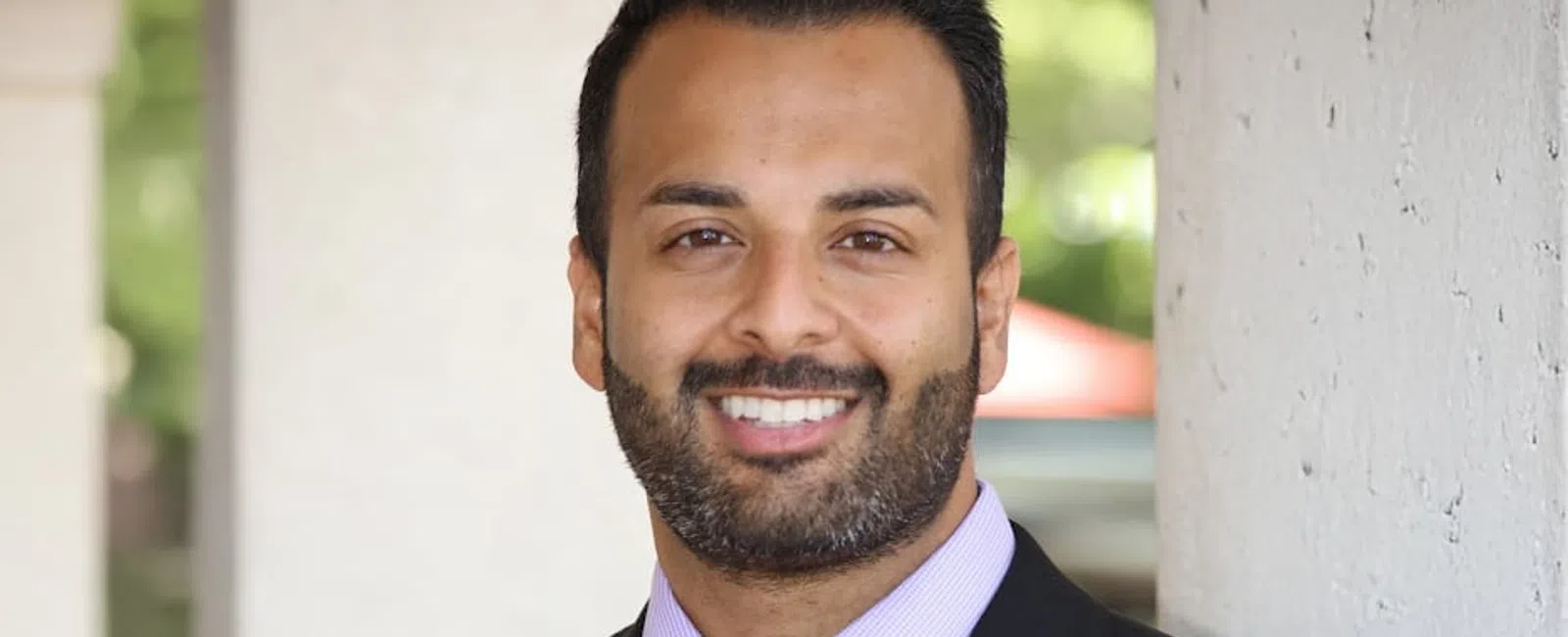 Smiling man in black suit and striped tie leaning against a wall with a blurred outdoor background.