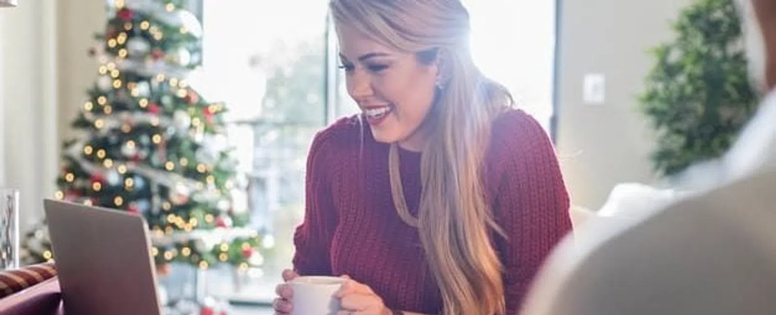Woman in red sweater smiling while video chatting on laptop near decorated Christmas tree with holiday lights.