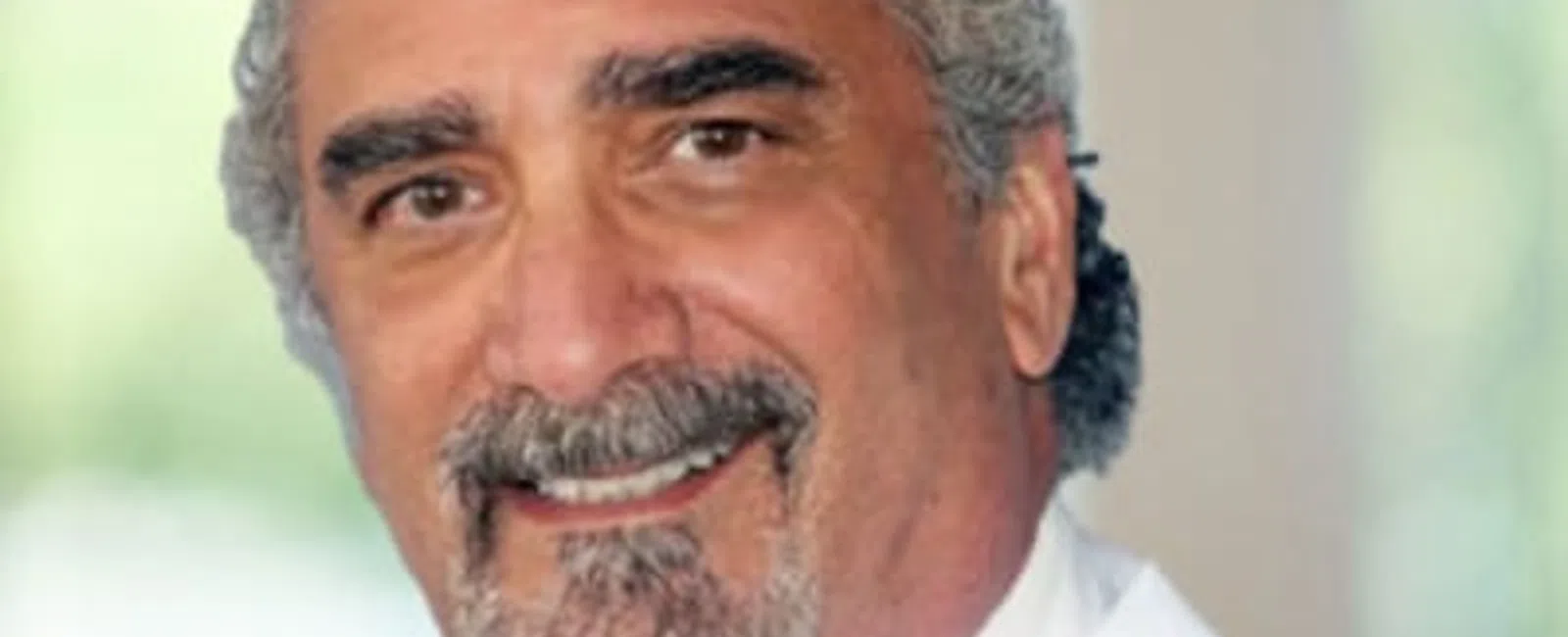 Smiling middle-aged man with gray hair and beard wearing white coat and patterned tie in a bright setting