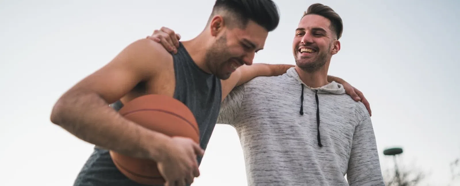 Two men outdoors smiling with one holding a basketball and the other with an arm around his shoulder.