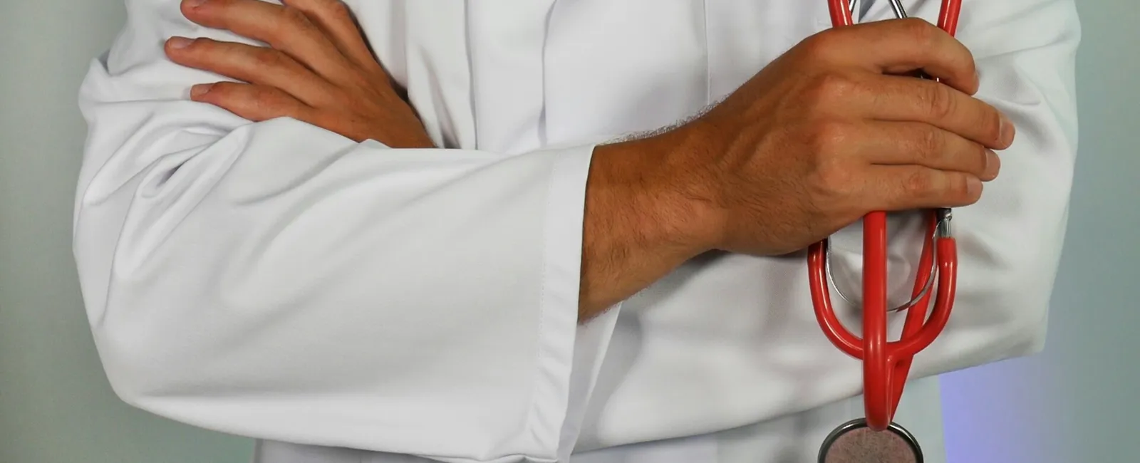 Doctor in white coat holding a red stethoscope with folded arms against a neutral background