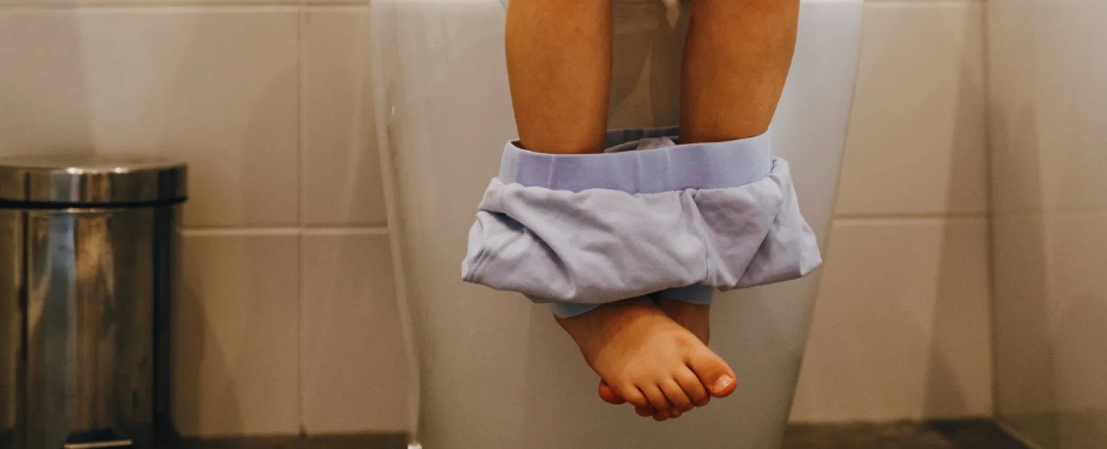 Child sitting on a toilet with pants down in a bathroom with tiled walls and floor.