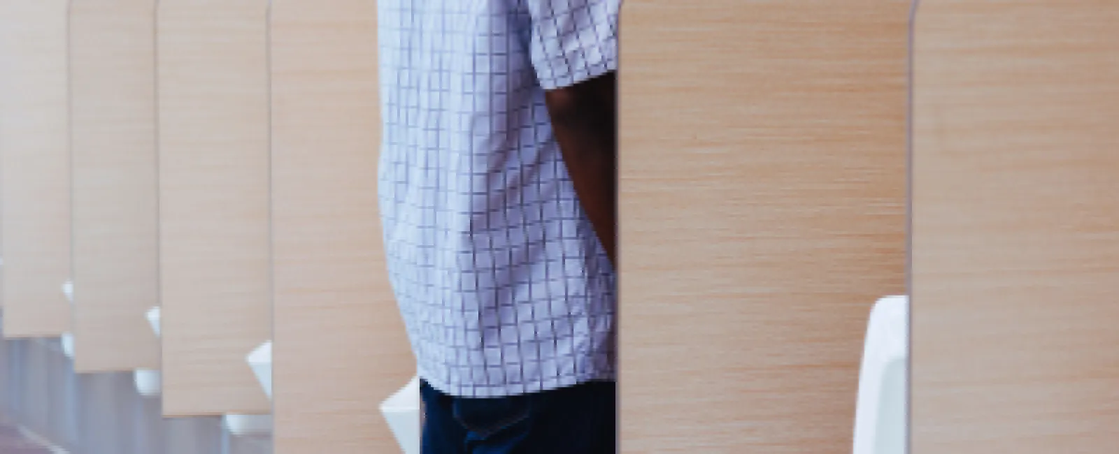 Man wearing a mask using a urinal with wooden privacy dividers in a public restroom.