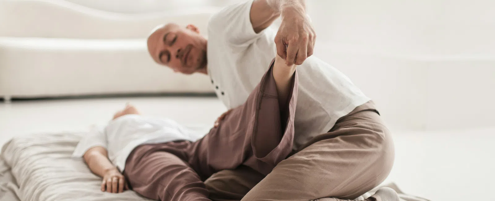 Man performing assisted leg stretch on woman lying on a mat in a bright room with a sofa and bed.