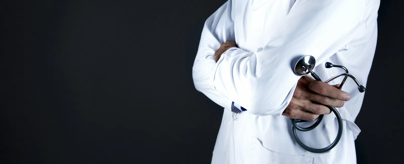 Doctor in white coat holding a stethoscope with arms crossed against a dark background