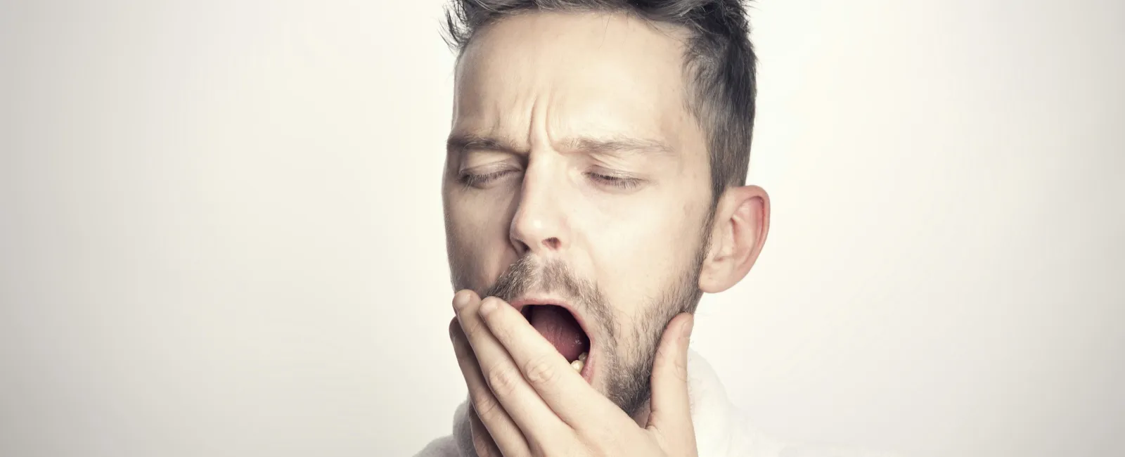 Young man with beard yawning and covering mouth, wearing a white robe against a neutral background
