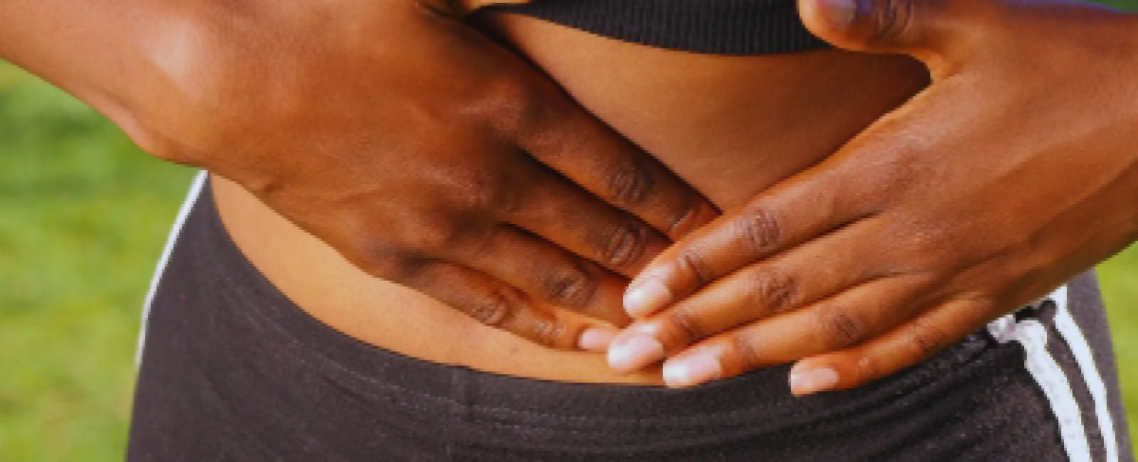 Woman wearing black workout clothes holding her stomach outdoors showing digestive discomfort.