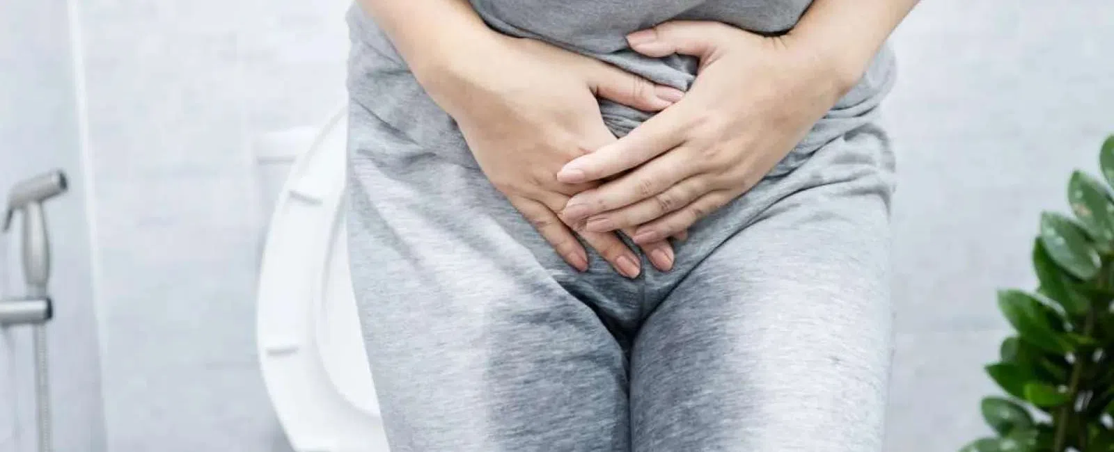 Person in gray clothes holding lower abdomen sitting on toilet, indicating urinary or bladder discomfort.