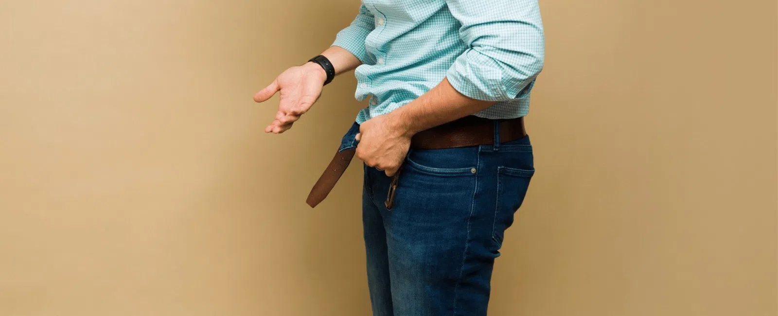 Man in glasses adjusting his belt wearing a blue checkered shirt and jeans against a beige background
