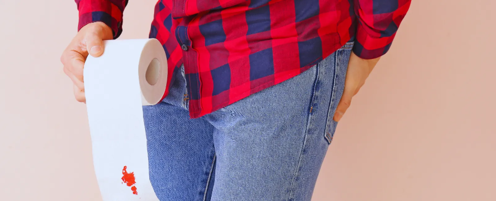 Person in red plaid shirt and jeans holding toilet paper with visible bloodstain on the paper.